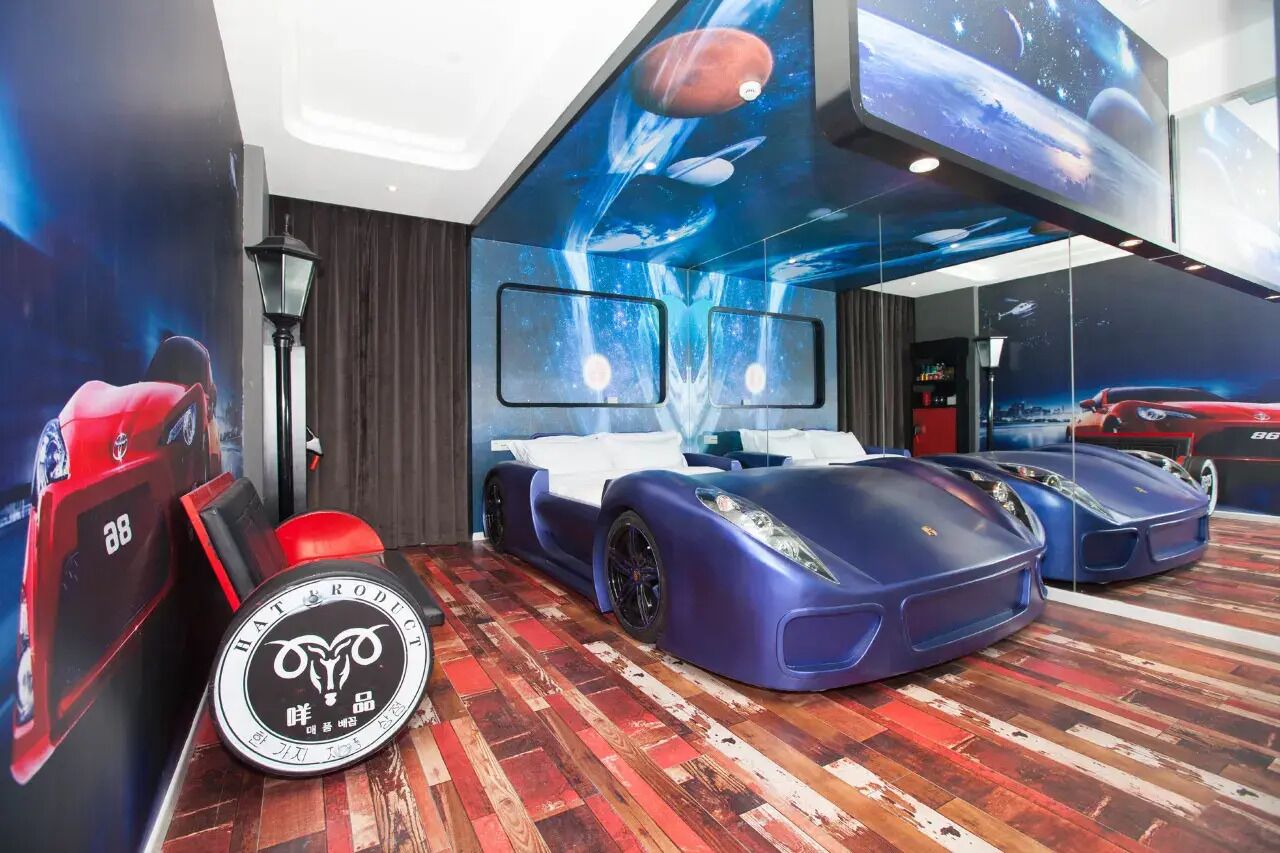 A room with a bed, a red sports car, and a blue sports car.