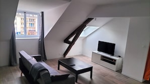 Eine moderne Loftwohnung mit schrägverdeckter Decke, einem grauen Sofa, einem schwarzen Couchtisch und einem Fernseher auf einem weißen Lowboard.