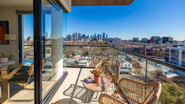 A balcony with wicker chairs and a table, overlooking a cityscape with modern buildings.