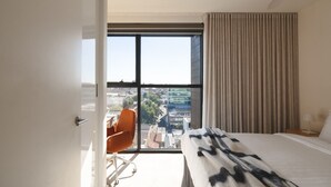 A hotel room with a large window, a bed with a patterned blanket, an orange chair, and a view of the cityscape.