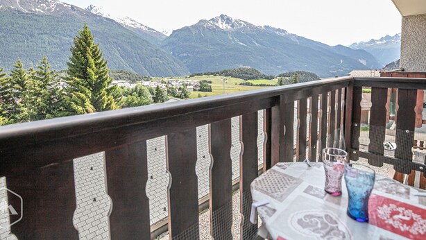 A balcony with a table and chairs, overlooking a mountainous landscape with snow-capped peaks and a village below. - Studio cabin 3 pers South balcony on snow front (Aussois)