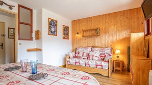 A cozy room with a wooden wall, a bed with patterned bedding, a small bedside table with a lamp, and a television mounted on the wall. - Studio cabin 3 pers South balcony on snow front (Aussois)