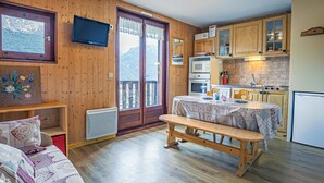 A cozy room with a wooden floor, a bench, a table with a tablecloth, a television mounted on the wall, and a kitchen area with wooden cabinets and appliances. - Studio cabin 3 pers South balcony on snow front (Aussois)