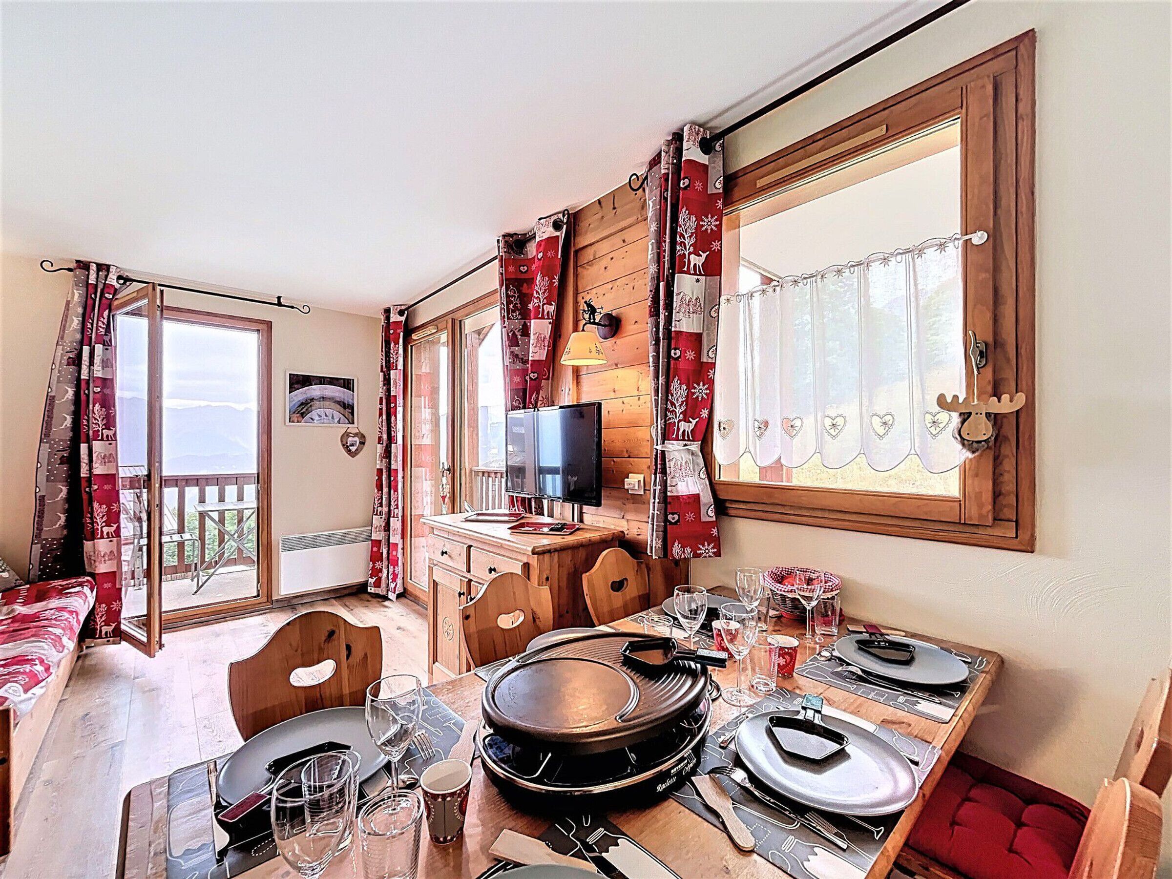 Een gezellig eetgedeelte met een houten tafel gedekt voor twee personen, een televisie op een houten standaard, en een balkon met rode gordijnen.