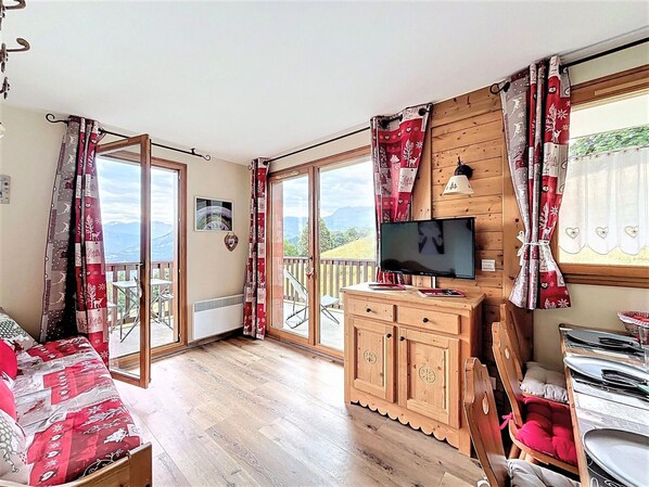A cozy room with a wooden bed, a TV on a wooden cabinet, and a balcony with a view of the mountains.