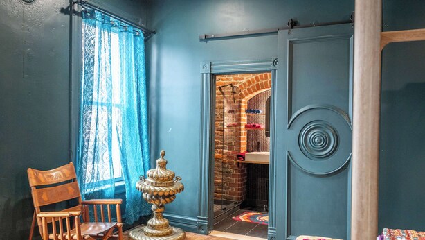 A room with a wooden rocking chair, a decorative floor lamp, and a door leading to a brick-walled bathroom. - Timeless on Main 2 For 2 (Blanchester)