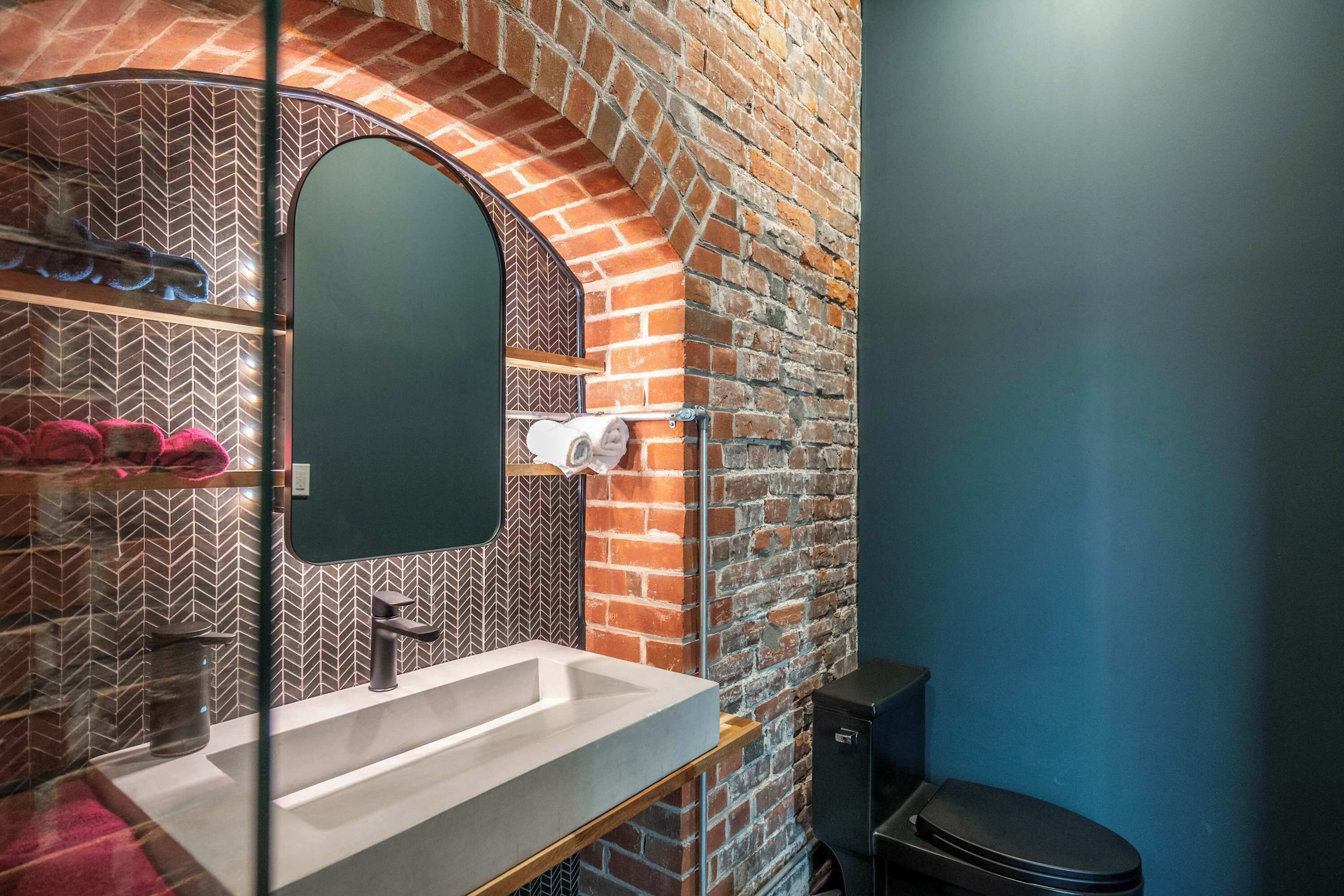 Une salle de bain avec un mur en briques, un lavabo blanc, un miroir et des toilettes noires.