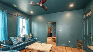 A room with a dark green wall, a ceiling fan, a wooden coffee table, a blue sofa with patterned cushions, and a wooden door. - Timeless on Main 2 For 2 (Blanchester)