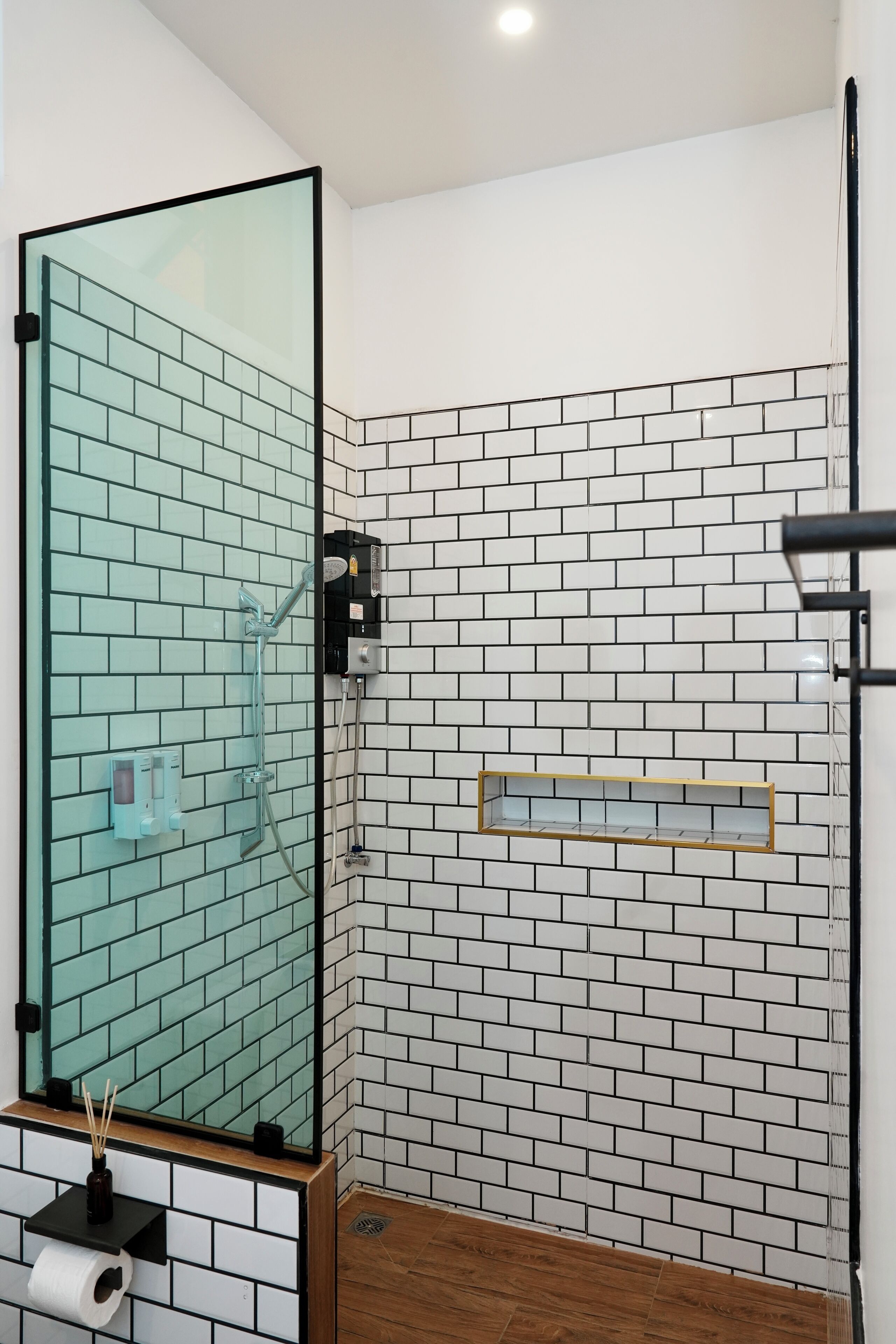Un baño con cabina de ducha de vidrio, paredes con azulejos blancos y negros, piso de madera y espejo con marco negro.