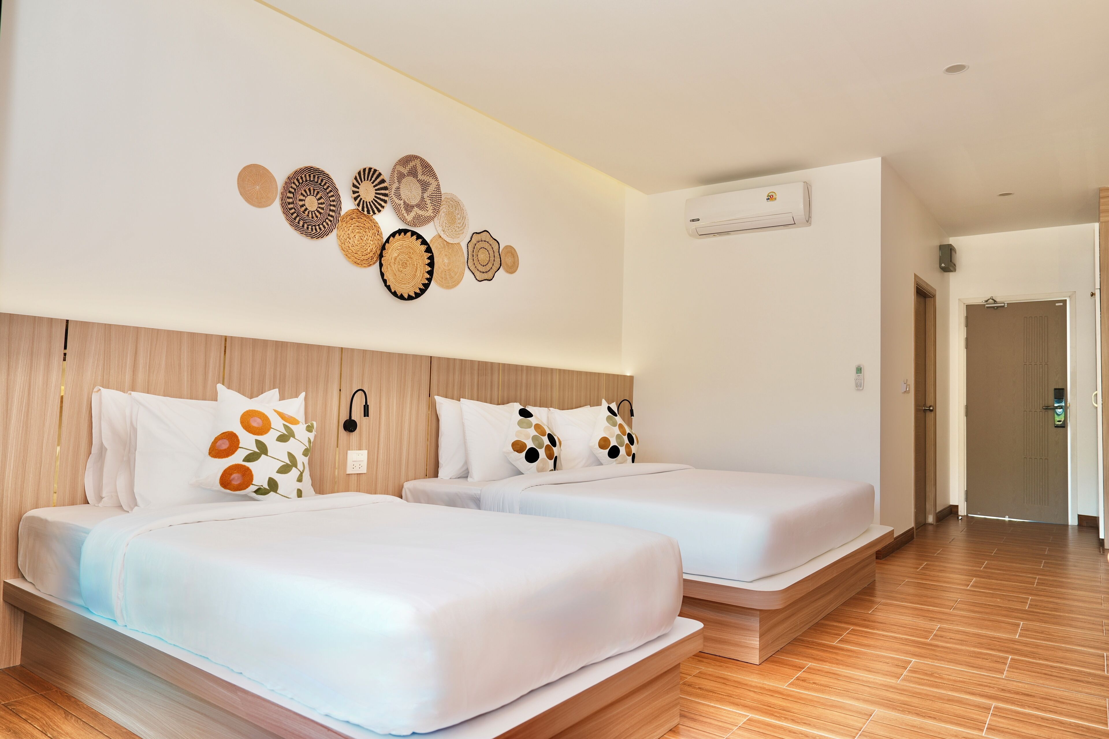 Quarto de hotel com duas camas, cabeceira de madeira, ar condicionado fixado na parede e um corredor que dá para outro quarto.