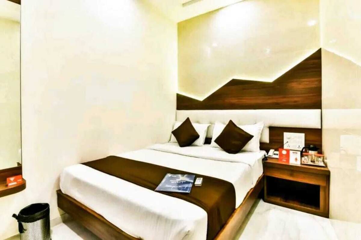 A hotel room with a neatly made bed, a wooden nightstand with a lamp, and a trash bin.