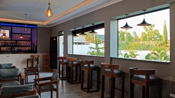 Un restaurante con sillas y mesas de madera, amplios ventanales con vistas a zonas verdes, iluminación colgante y una zona de bar con estantes llenos de botellas.