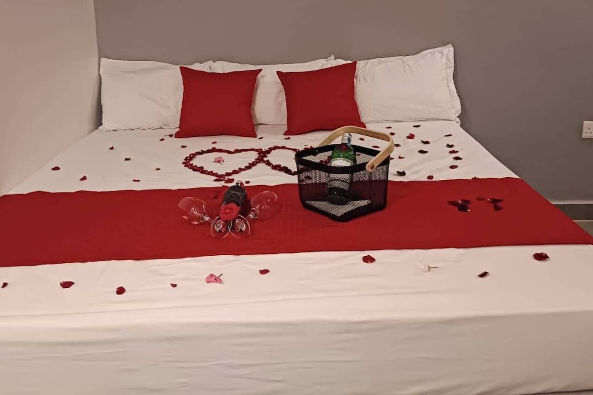 A bed with white and red bedding, a basket with a bottle, and rose petals scattered on the bed.