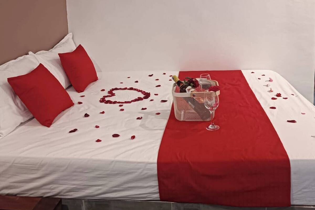 A bed with red and white bedding, a heart-shaped arrangement of rose petals, and a basket with a bottle and glasses on the side.