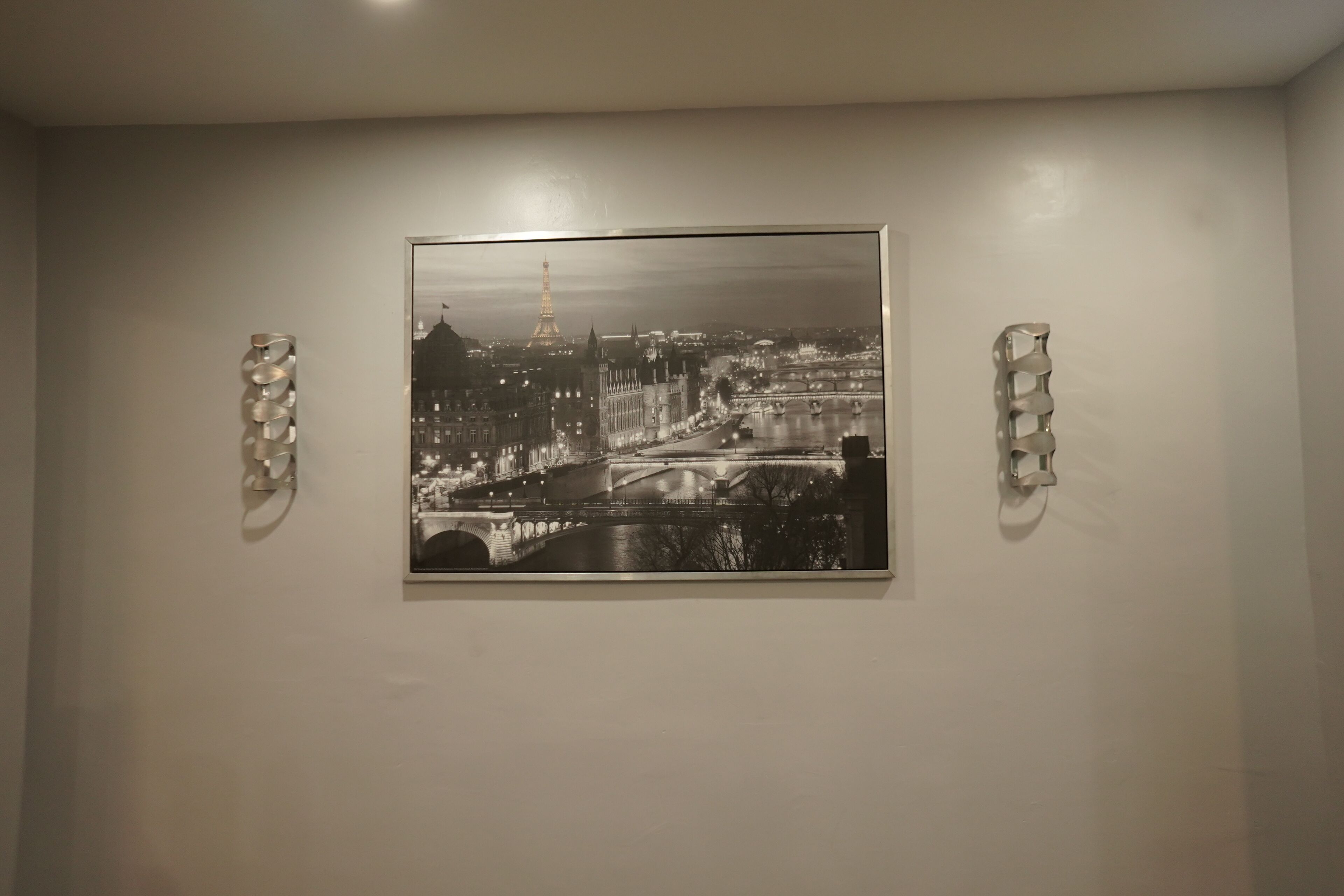 A framed black and white photograph of a cityscape with a bridge and the Eiffel Tower, mounted on a plain wall with two abstract wall sconces.