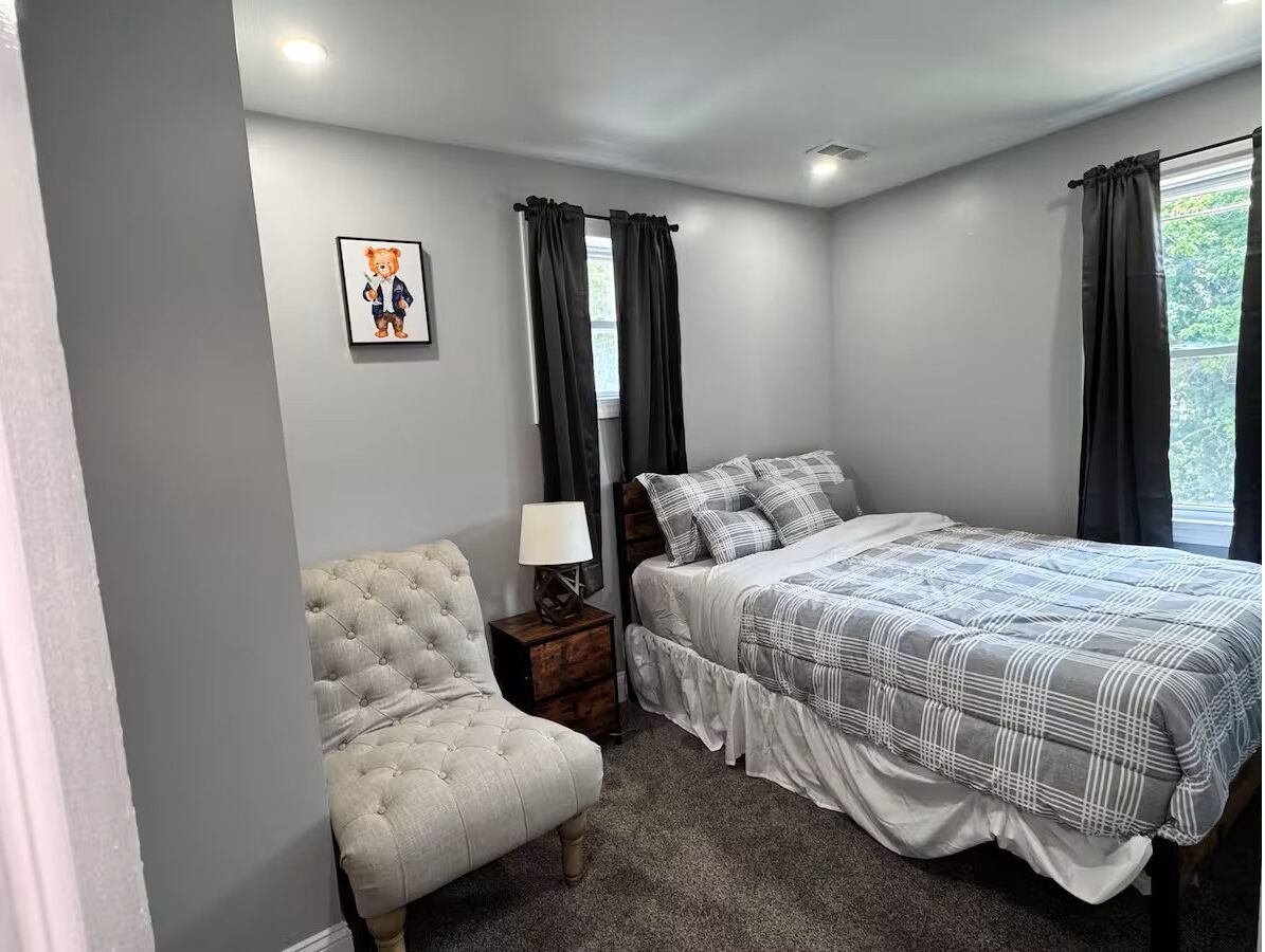 A bedroom with a bed, a chair, a nightstand with a lamp, and a picture on the wall.