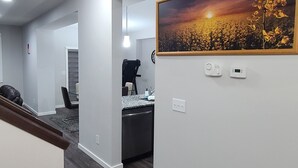 Una cocina moderna con horno integrado, una zona de comedor con una silla, y un pasillo que conduce a un salón con un cuadro grande.