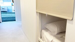 A small, neatly arranged room with a bed, a door, and a view of a blue outdoor area. - Single capsule in room of 16 capsules (El Palmar de Vejer)