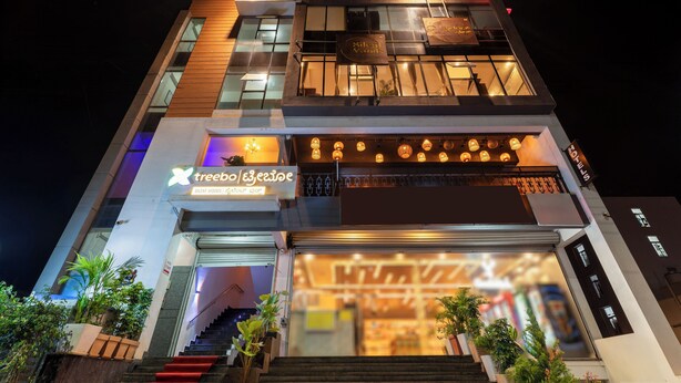 A multi-story hotel with a modern design, featuring a staircase leading to the entrance, illuminated signage, and potted plants at the base. - Treebo Silent Woods (Bengaluru)