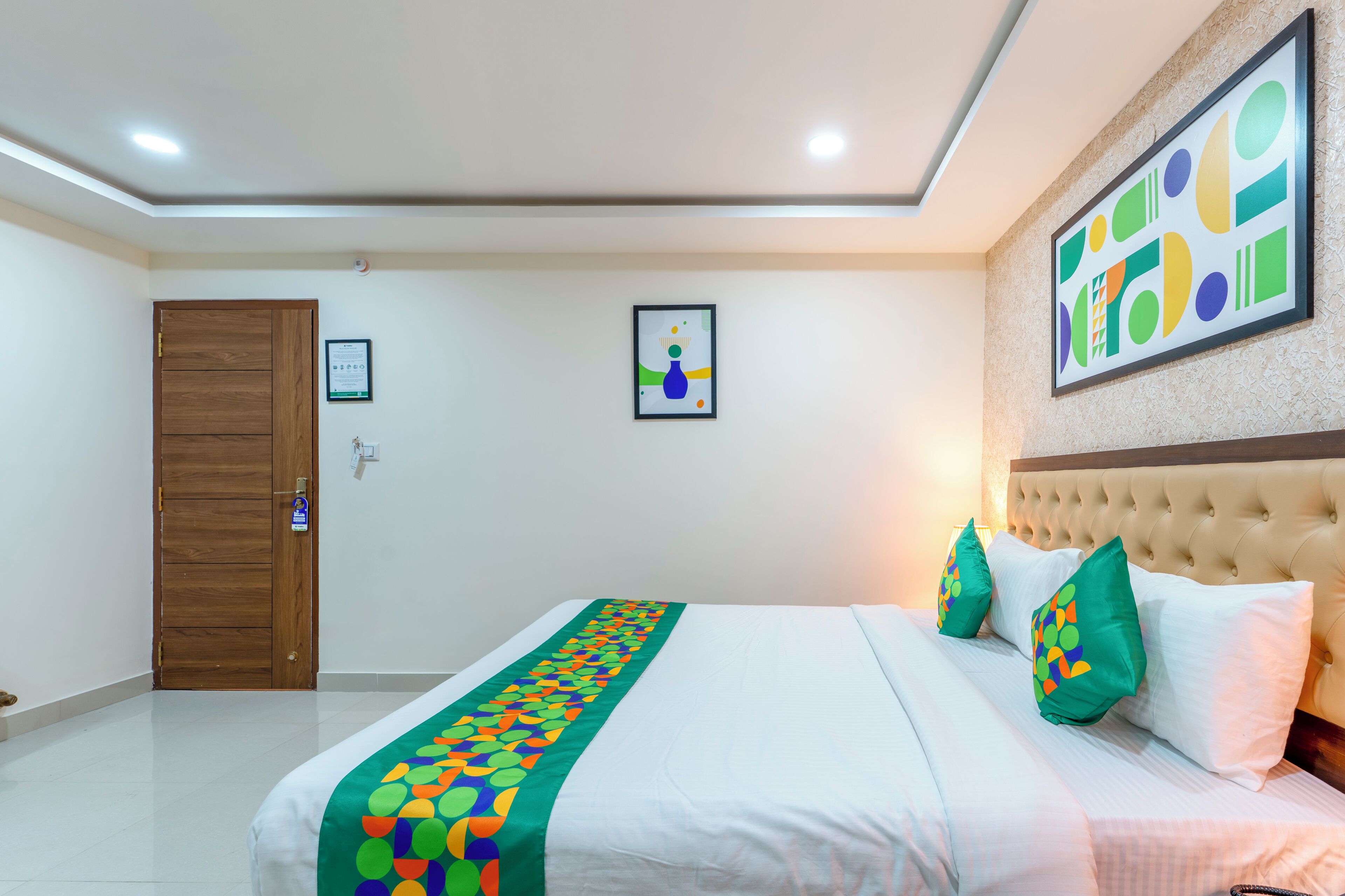 A hotel room with a bed, a wooden door, a framed picture on the wall, and a ceiling light.