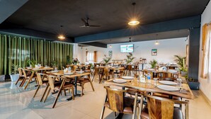A spacious dining area with multiple wooden tables and chairs, ceiling fans, and potted plants. - Treebo Silent Woods (Bengaluru)