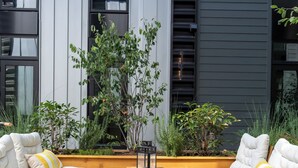 Zona d'estar a l'exterior amb dos sofàs blancs amb coixins, una taula de fusta i jardineres amb diferents plantes.