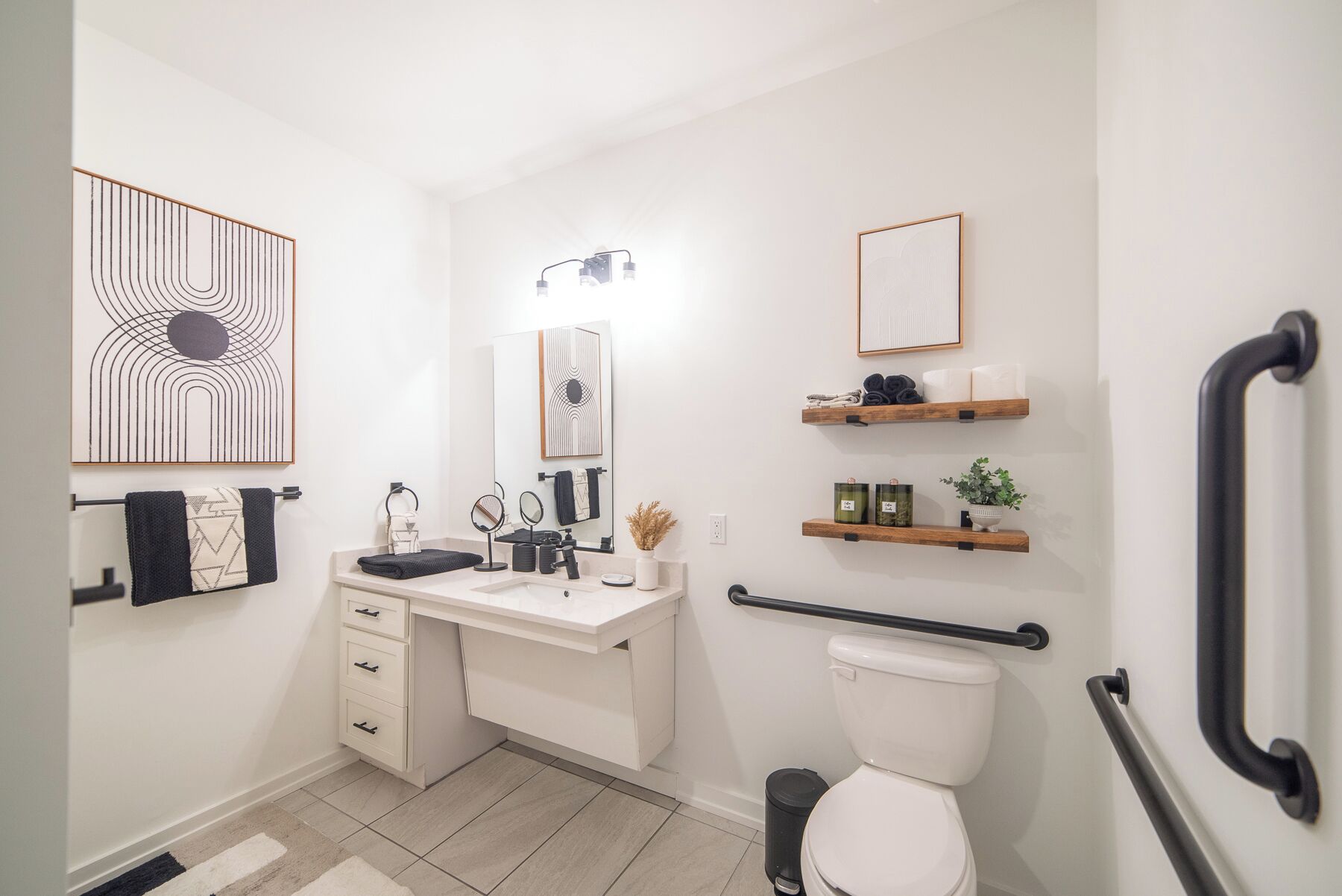 A modern bathroom with a white vanity, a toilet, and a shower area. There are shelves with toiletries and a handrail for accessibility.