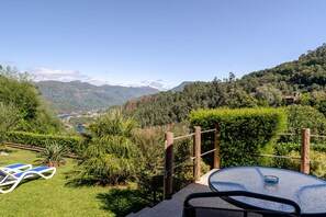 A patio with a table and chairs, surrounded by lush greenery and a mountain view.