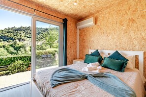 A bedroom with a large window, a bed with pillows, a bedside table, and a view of a green landscape.