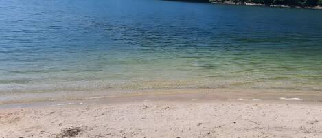 A beach with clear water, sandy shore, and forested hills in the background.
