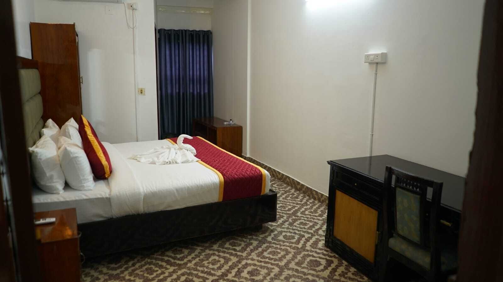 A hotel room with a bed, a wooden nightstand, a television, and a patterned carpet.