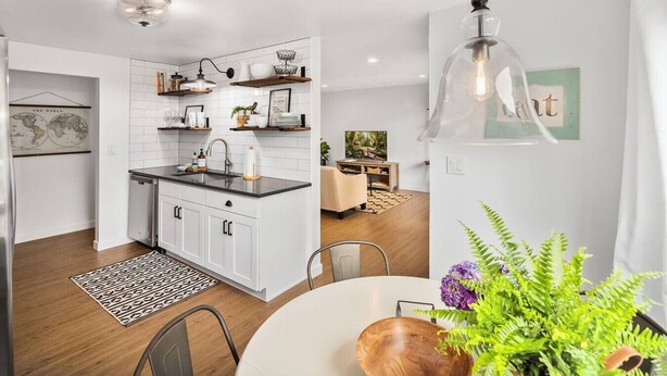 Een moderne keuken met witte kasten, een zwart aanrecht en een hanglamp. Er is een eethoek met een ronde tafel en stoelen, en een plant op de tafel.