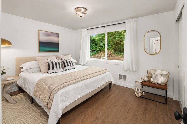 A bedroom with a bed, a bedside table, a lamp, a window with curtains, a mirror, and a view of greenery outside.