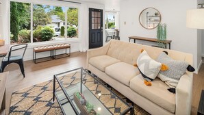 Een moderne woonkamer met een beige bank, een glazen salontafel en uitzicht op een tuin door grote ramen.