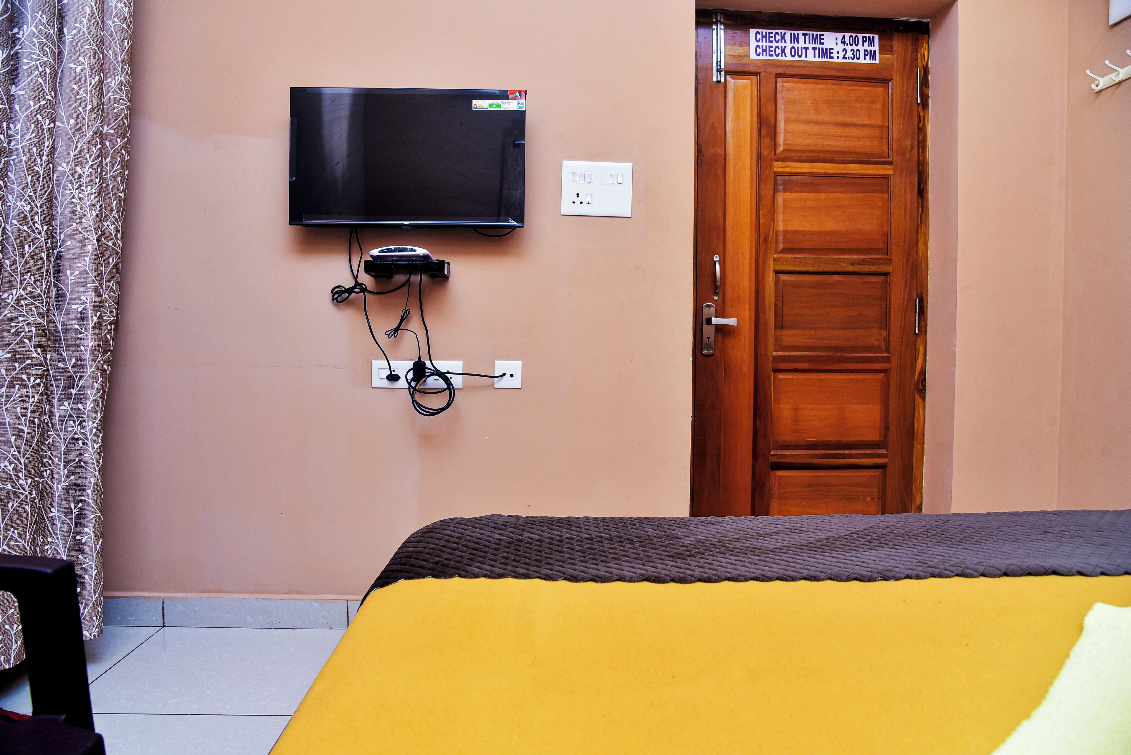 Een hotelkamer met een bed, een aan de muur bevestigde televisie, een deur met een bord waarop in- en uitchecktijden staan, en een stopcontact.