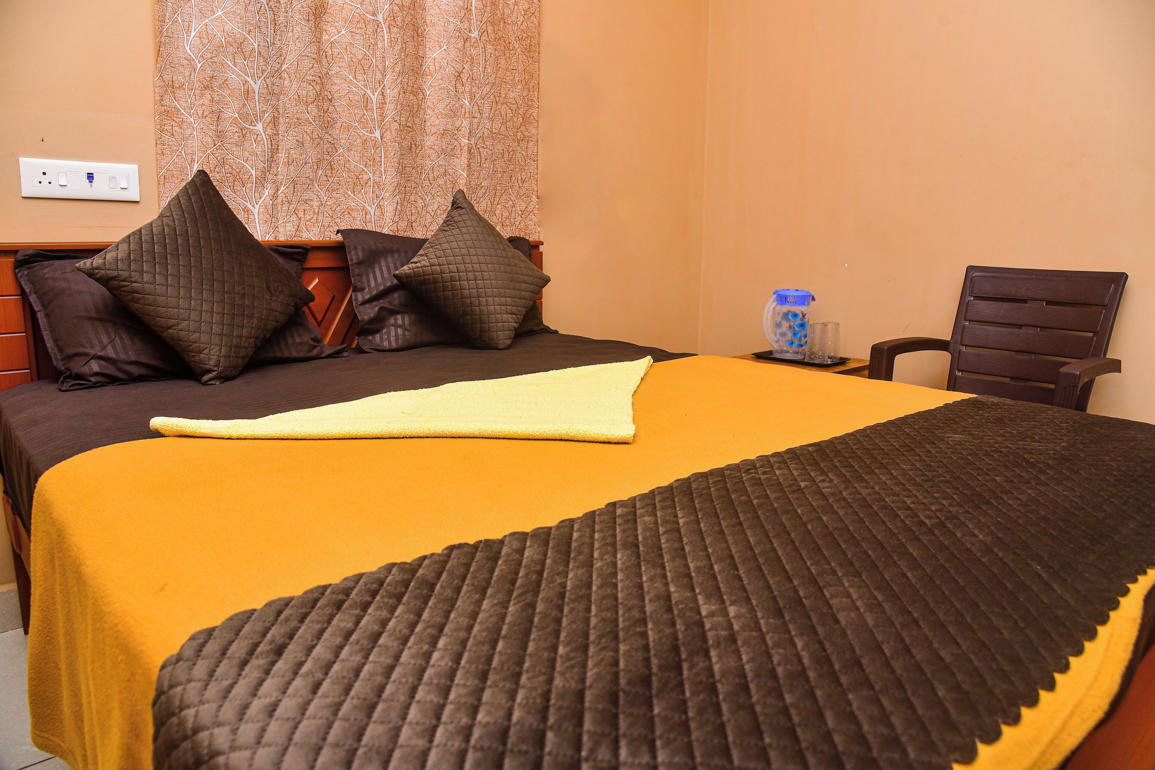 A neatly made bed with a yellow and brown bedspread, a brown quilted blanket, and two pillows. There is a wooden nightstand with a blue water bottle and glasses beside the bed. A brown chair is positioned against the wall.