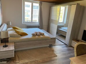A bedroom with a bed, bedside table, lamp, and a view of the outside through a window. - BLISS Basel Residence (Binningen)