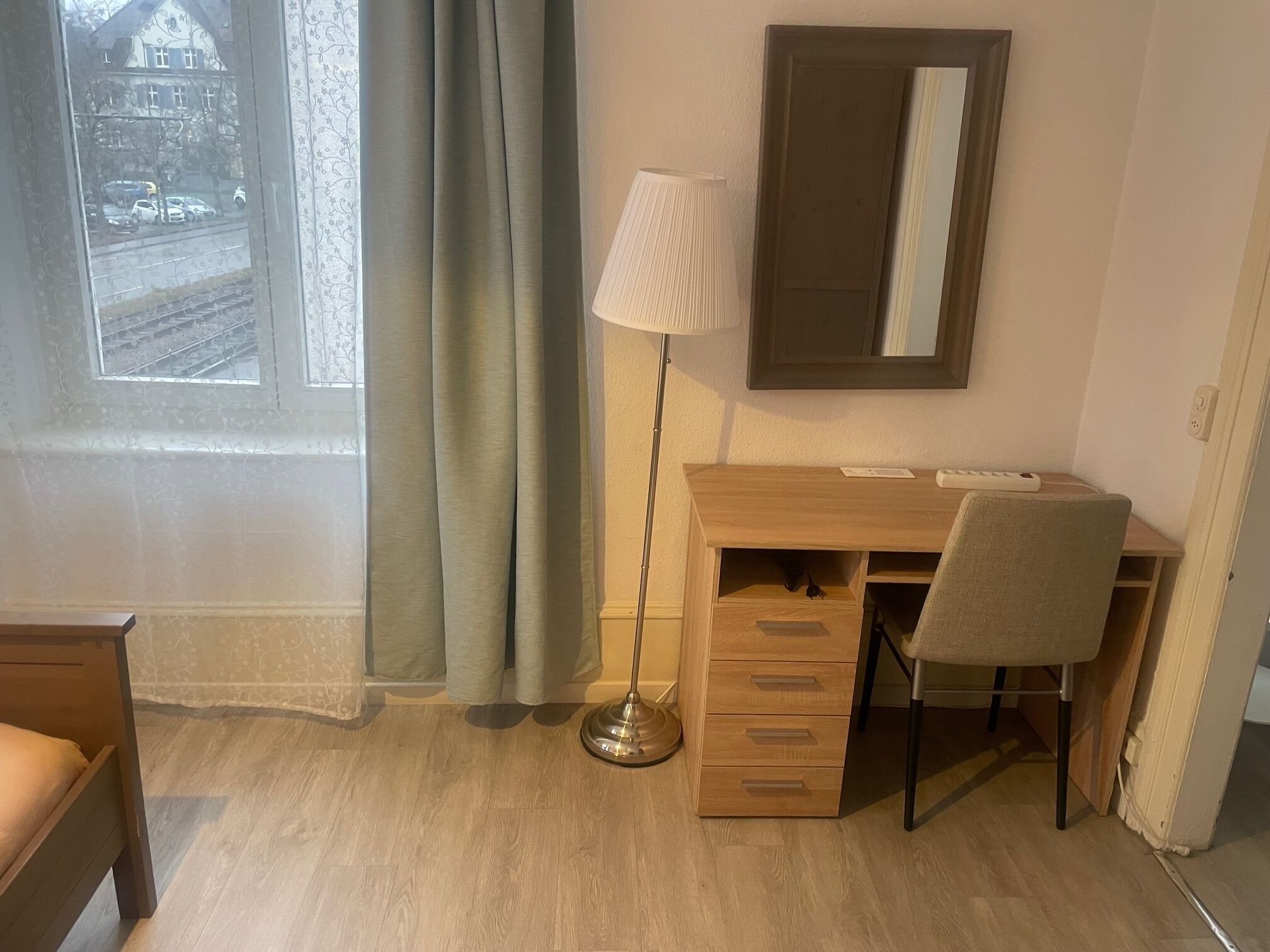 A room with a wooden desk, a chair, a lamp, a mirror, and a bed.