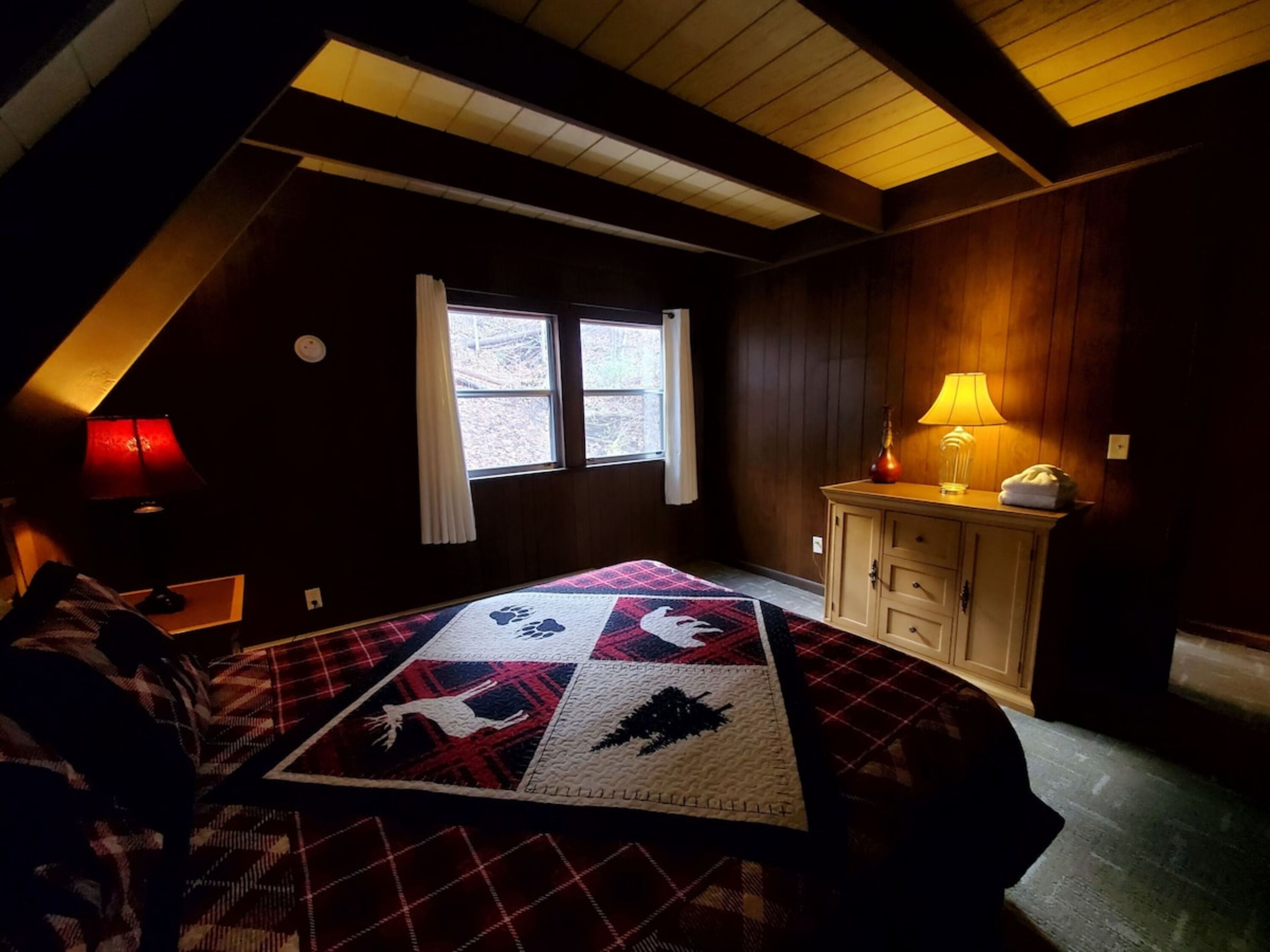 A cozy attic bedroom with a sloped ceiling, a large window, a quilt on the bed, and a bedside lamp.