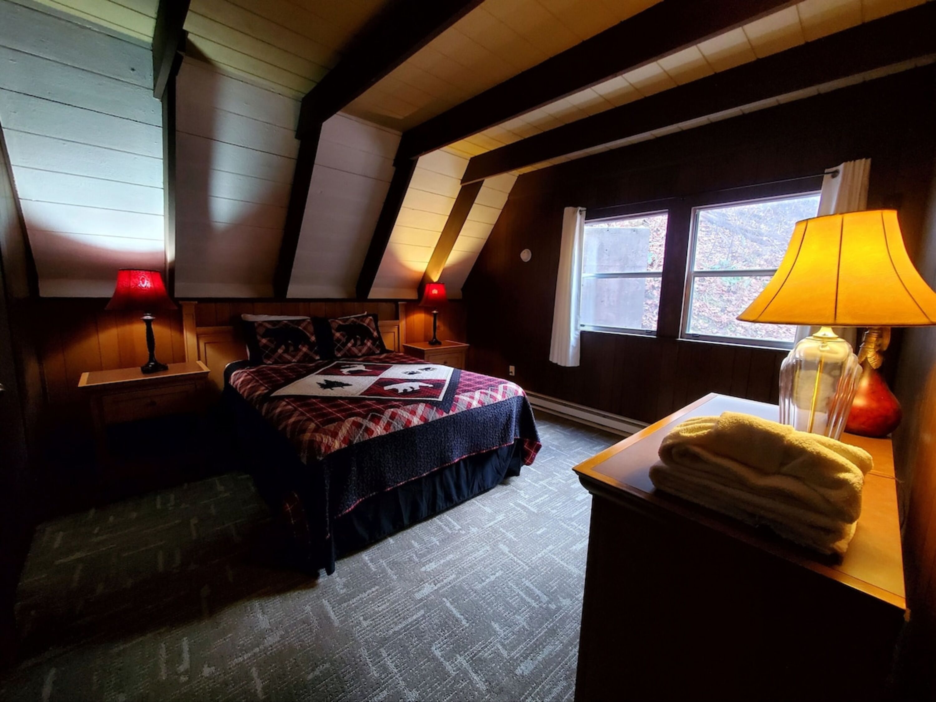 Een gezellig zolderkamer met een schuine kap, een bed met quilt, twee bedlampen, een raam met uitzicht en een nachtkastje met lamp.