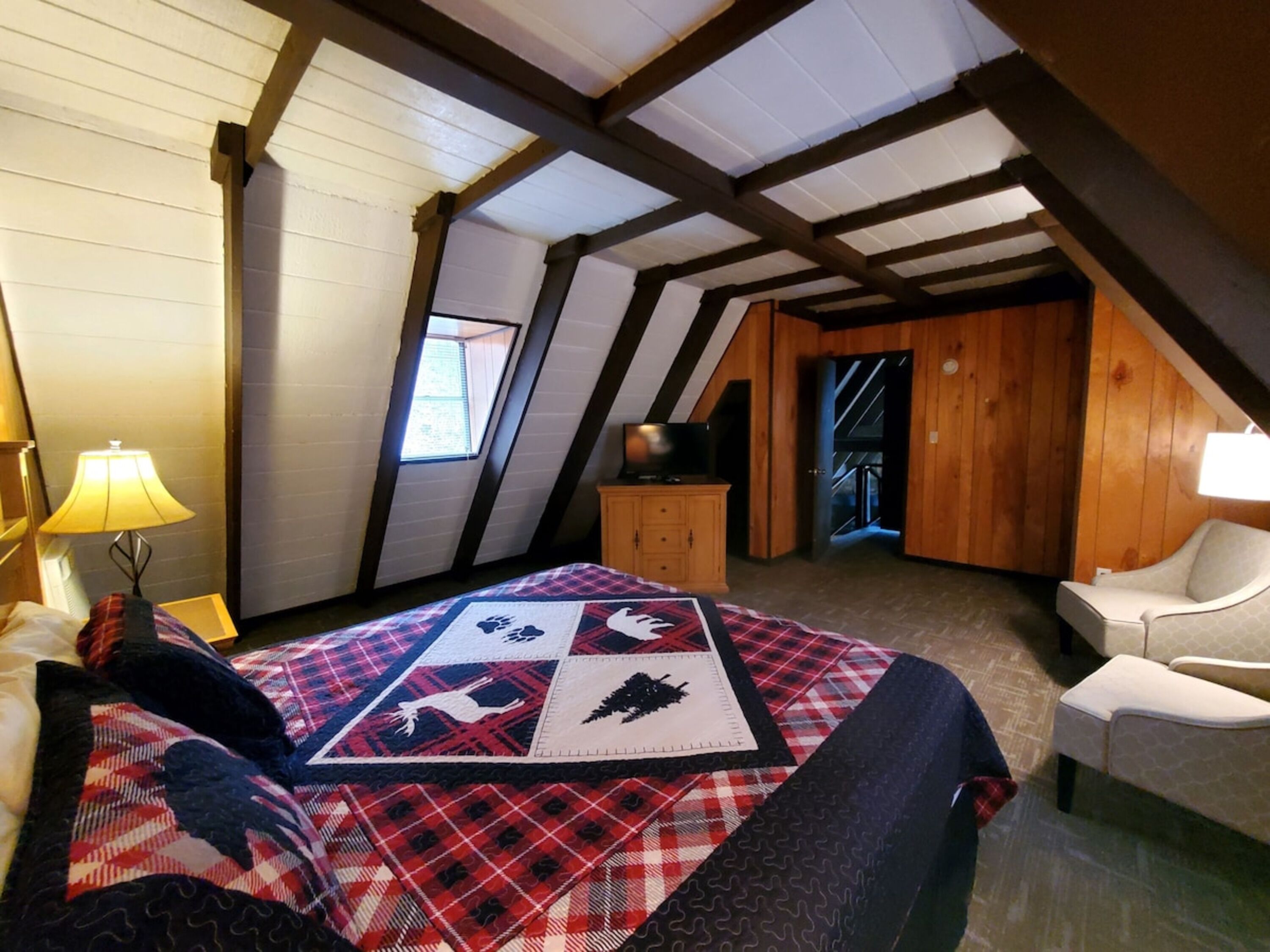 Une chambre mansardée chaleureuse, avec un plafond en pente, un lit recouvert d’une couverture à carreaux, une chaise et une petite armoire en bois.
