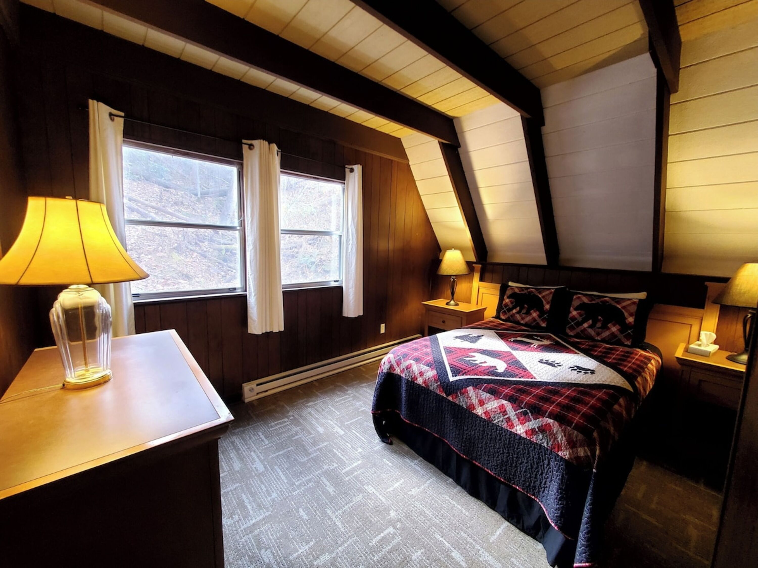 Une chambre confortable avec un plafond en bois, un lit recouvert d’une courtepointe à motifs, deux tables de chevet avec des lampes et une fenêtre avec des rideaux.
