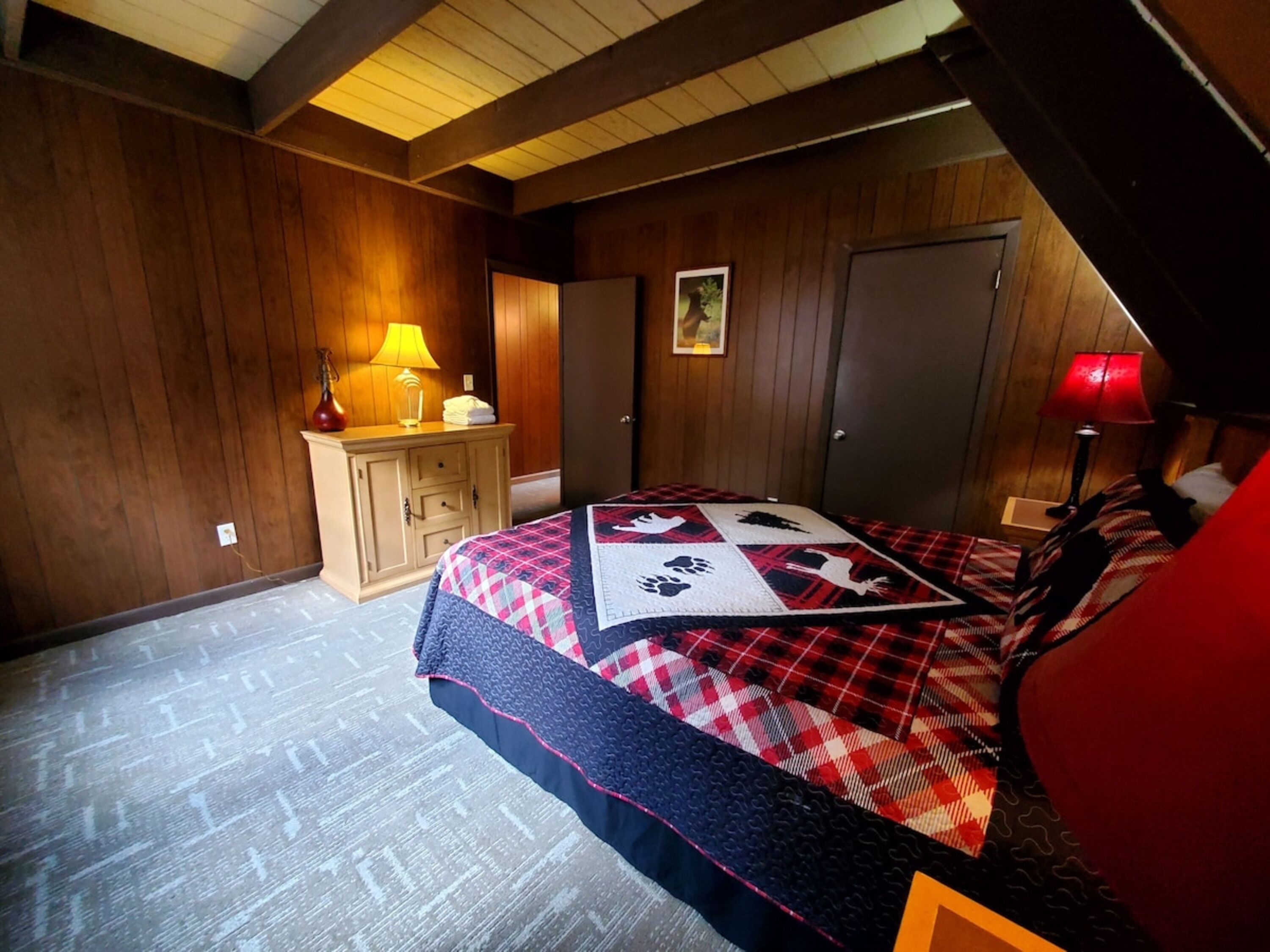 Un dormitorio con paredes de madera, una cama con colcha escocesa, una mesita de noche con lámpara y un aparador con lámpara.