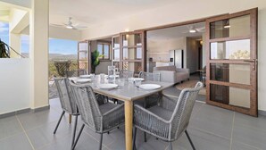 A dining area with a table set for four, surrounded by wicker chairs, and a view of a kitchen and living area through glass doors.