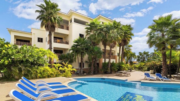 A pool area with lounge chairs, palm trees, and a building with balconies.