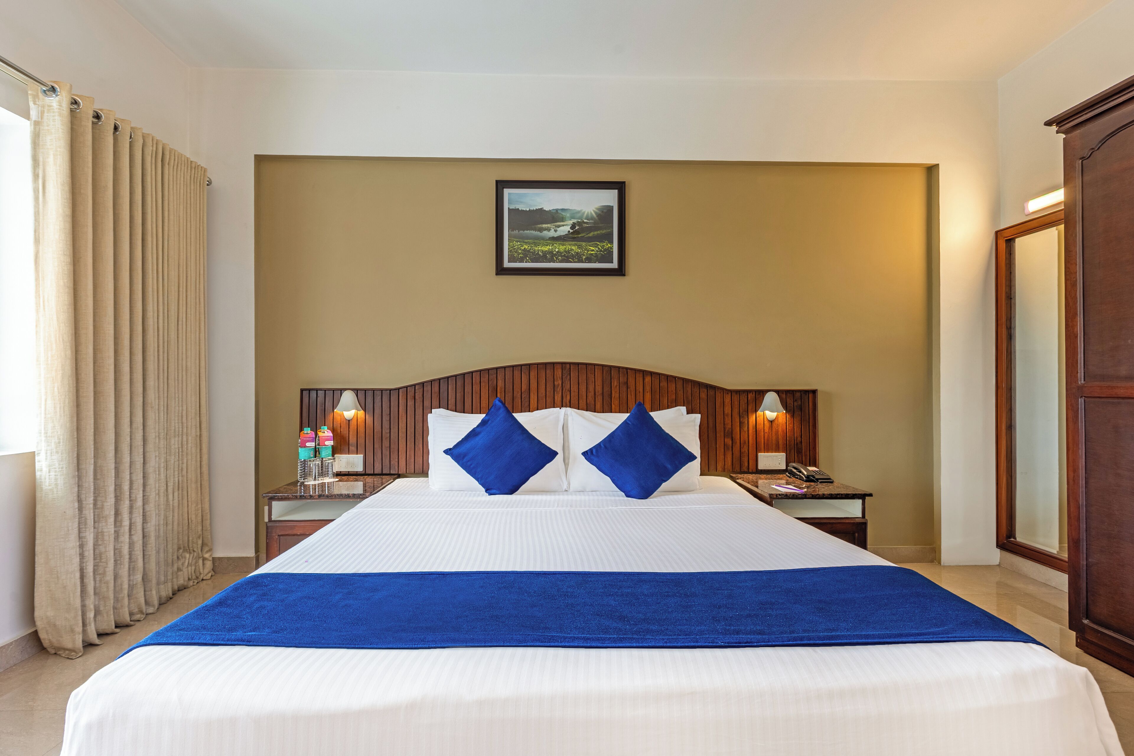 A hotel room with a large bed, two nightstands, a wooden headboard, a framed picture on the wall, and a window with curtains.
