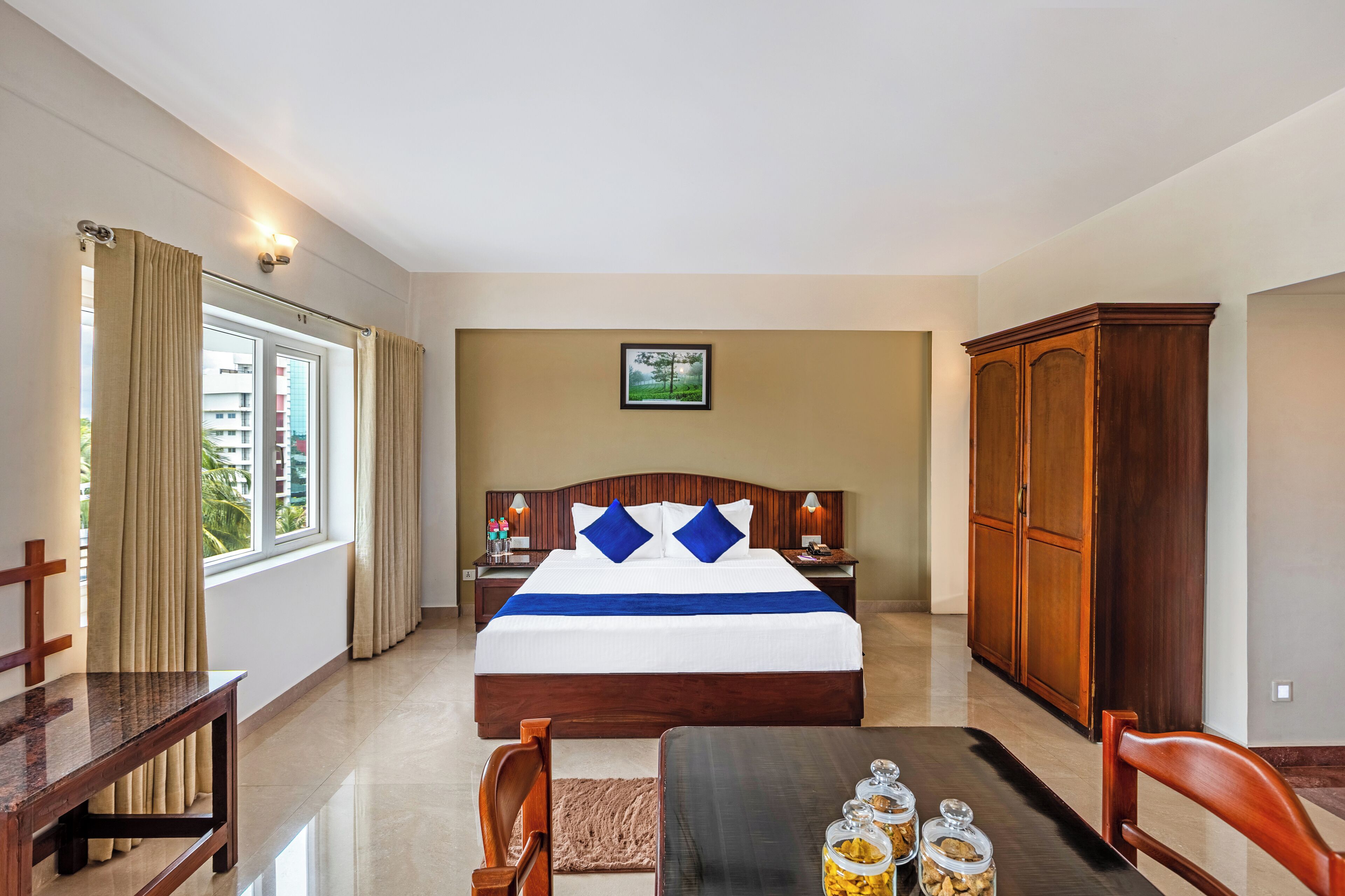 Une chambre d’hôtel avec un grand lit, des meubles en bois, une table à manger avec des bocaux et une vue sur les bâtiments par la fenêtre.