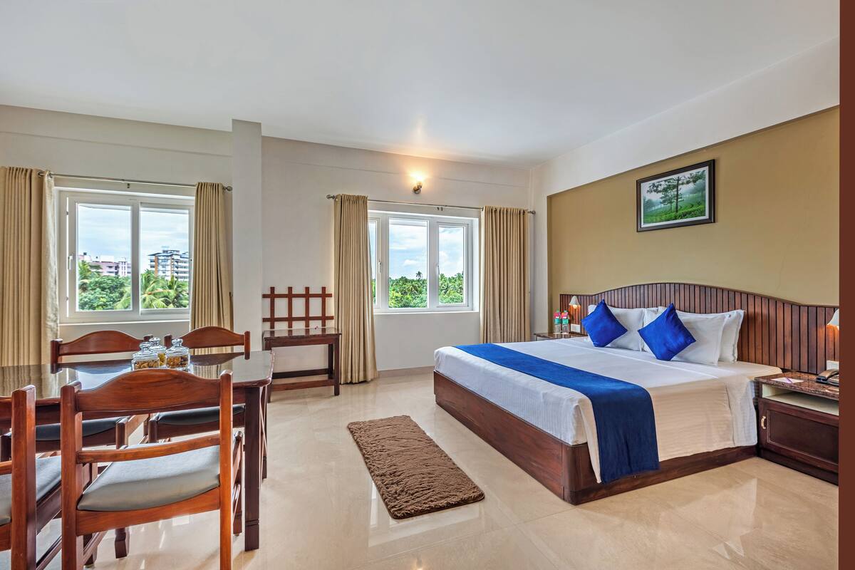 Habitación de hotel con una cama grande, muebles de madera, una mesa de comedor y vistas a espacios verdes.
