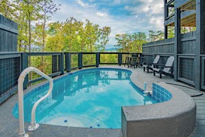 Une piscine dotée d’une terrasse en bois, entourée d’une rambarde en métal, et offrant une vue sur une zone boisée.