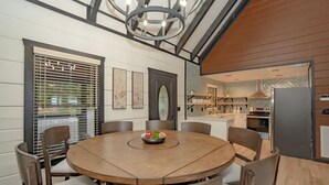 A dining area with a round wooden table and chairs, a chandelier, and a view of the kitchen through a window with blinds.